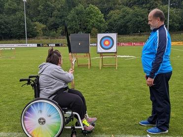 Foto zeigt ein Mädchen im Rollstuhl, welches Bogen schießt. Der Trainer steht nebendran